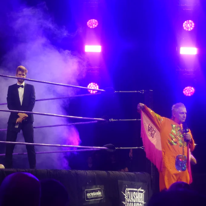 Joe Lycett lifting up an arm to reveal LEFT WING written above the pink fringing, as Ivo Graham stands above him in the ring, in a smoky haze