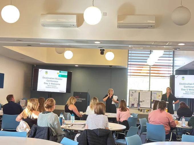 Two speakers standing in front of a room of people sitting at tables. Two screens are in the background that say 'Destination Reader Conference Day 1 Welcome'