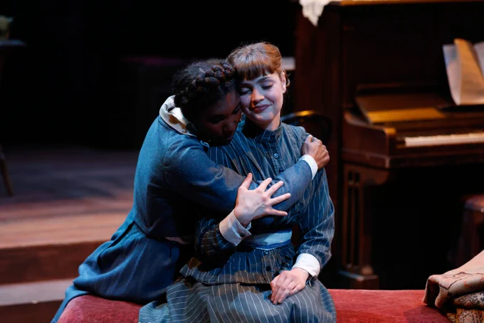 Two actors embrace warmly on stage in a scene from a theater production, with a piano in the background.