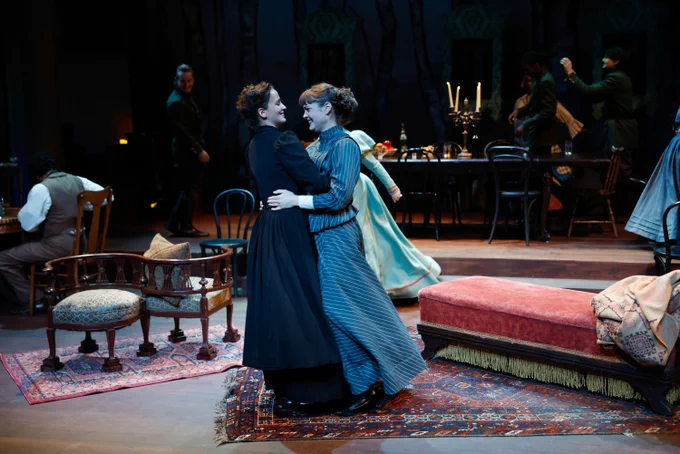 Two actors portraying a heartfelt embrace on a stage set decorated in a Victorian style, with other performers in the background, during a theatrical production.