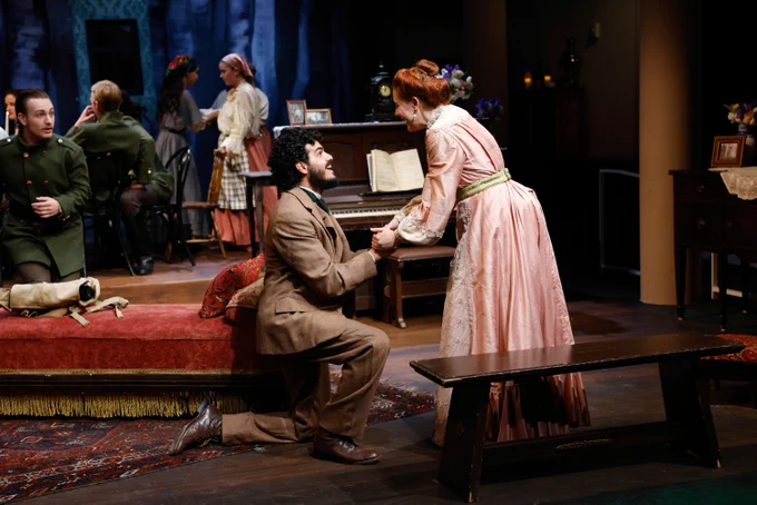 A scene from a theatrical production showing one actor kneeling and holding the hand of another actor who is standing, both in period costumes, on a stage set designed to resemble a cozy, historical living room. Other actors are in the background, also in period attire.