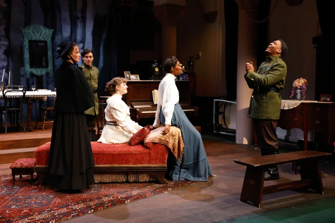 A theatrical scene with five actors on a vintage-style set, featuring intricate decorations, a piano, and period-appropriate furniture. The actors are dressed in early 20th-century costumes, engaging in a dramatic conversation with varying expressions of intrigue and delight.
