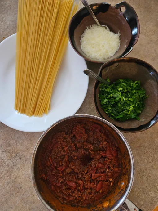Ingredients clockwise from left of a soup plate with a handful of dry spaghetti, a bowl of grated cheese, a bowl of chopped parsley, a small pot meat sauce.