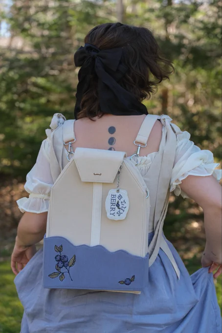 Woman standing in front of a Forrest wearing a blue skirt and a white puff sleeve chemise dress with a backpack on her back shaped like a giant teabag. The bottom of the teabag is blue, blueberry tea, and the tag hanging off of the teabag says blueberry tea.