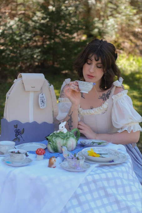 A girl with curly brown medium length hair is sitting at a small table, wearing a white puff sleeved dress and blue skirt. The table is covered in a white and gray checkered tablecloth and set for a tea party. On the table is a large purse in the shape of a teabag. She’s holding a small cup of tea about to take a sip.