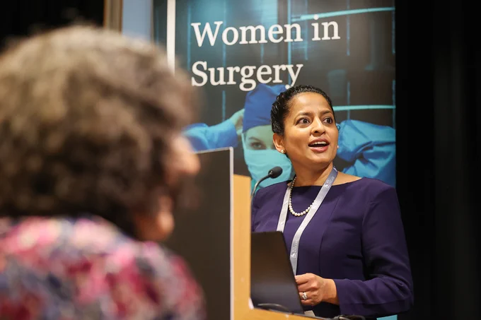 A woman in a purple dress and pearl necklace speaks at a podium with a 'Women in Surgery' banner behind her.
