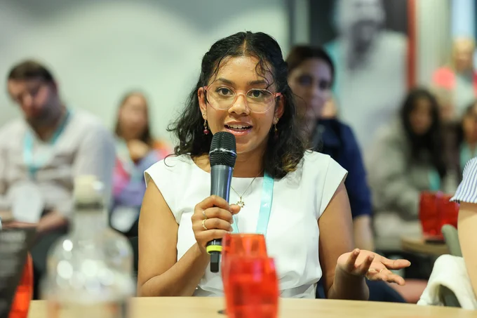 Woman in white sleeveless top holding a microphone and speaking during a group discussion or event.