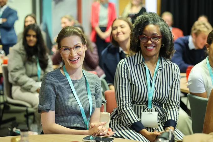 Two women wearing conference badges sit attentively at a round table during a seminar or workshop.