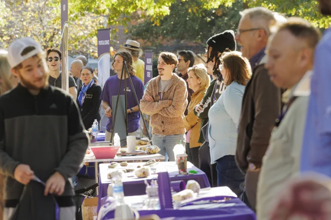 Group of people attending ECU's Open House outdoor event, standing near a table covered with displays under trees with autumn leaves.
