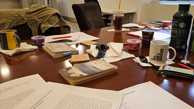 Desk covered with books, papers, pens, and breakfast snacks
