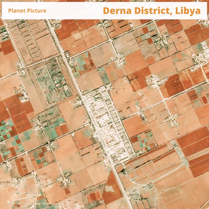 This satellite image shows a section of the Derna District in Libya, characterized by a patchwork of agricultural fields and rural settlements. The land is divided into rectangular plots in varying shades of reddish-brown and green, indicating different stages of cultivation and vegetation cover. A main road runs vertically through the center, lined with clusters of buildings that form small towns or farming communities. The landscape reflects a semi-arid environment where agriculture depends heavily on irrigation and seasonal rainfall.