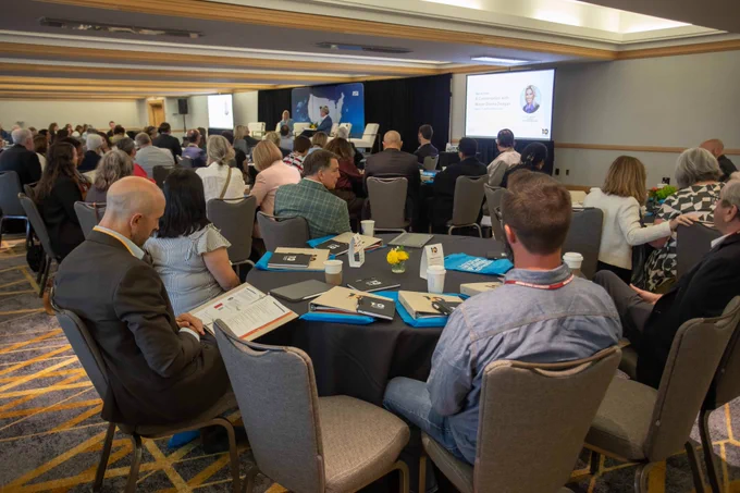 a large room of people seated at tables listening to a speaker at the front of the room at the ten across convergence meeting