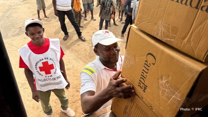 Mozambique Red Cross workers smile as they unload cardboard boxes marked with the Government of Canada logo from a truck. The boxes, provided by Canada and the Canadian Red Cross, contain critical relief items for communities affected by Cyclone Chido. Civilians, including children, can be seen in the background.
