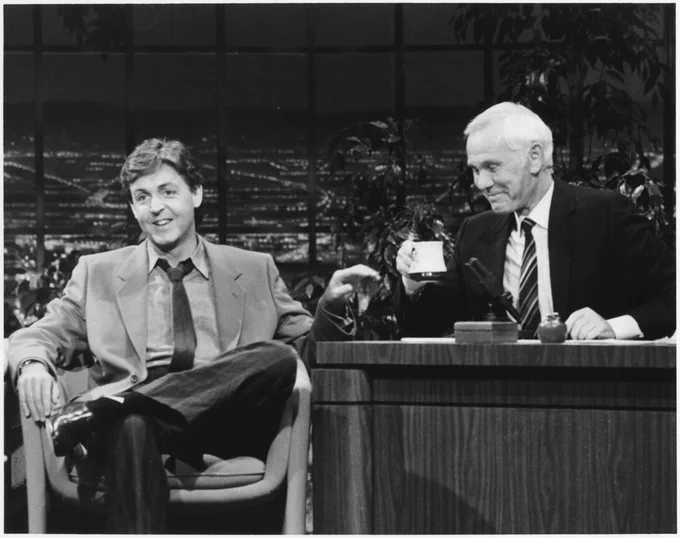 Johnny Carson interviews musician Paul McCartney on "The Tonight Show" in this black-and-white photo. Carson (right), seated behind a desk, is wearing a dark suit coat, white shirt and diagonally striped tie. He closes his eyes and smiles as he  holds up a coffee mug. McCartney (left), seated in a guest chair, is wearing a lighter-colored suit and dark tie. He smiles as he looks toward the studio audience. (Courtesy)
