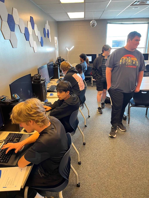High school students working on computers. Two students walking behind them.