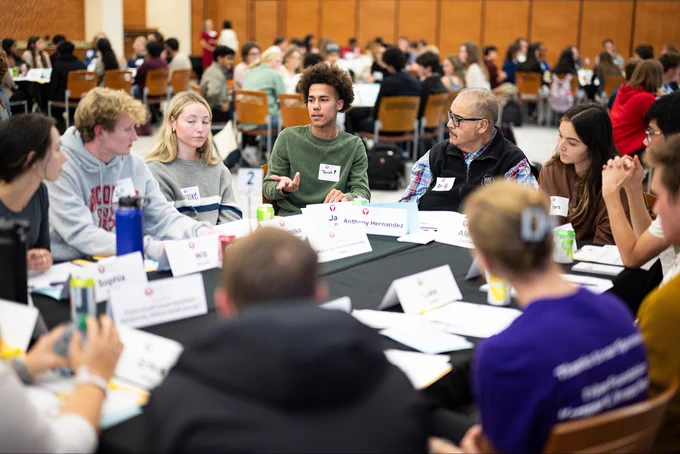Several students are engaged in conversation around a table at a Deliberation Dinner with name tags and some students wearing school apparel.