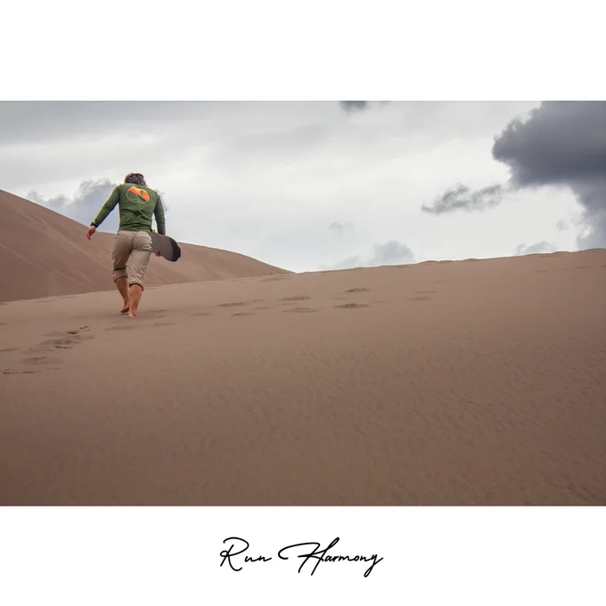 This striking photograph was taken at Great Sand Dunes National Park and Preserve in Colorado, capturing a moment of solitude and adventure beneath a dramatic sky. A person climbs barefoot up the vast, wind-sculpted dunes, carrying a sandboard in hand. The smooth, textured surface of the sand contrasts beautifully with the moody clouds above, evoking a sense of scale, perseverance, and wonder. The muted earth tones and soft lighting emphasize the quiet grandeur of the landscape—where endless dunes meet the ever-changing sky. This image embodies both the physical challenge and meditative calm of exploring one of America’s most unique natural environments, where nature’s simplicity transforms into art through motion, light, and perspective.
