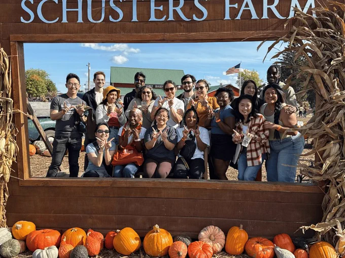 A group of 17 ELPA MS and PhD students smile and make the UW–Madison "W" with their hands in a large wooden frame at Schuster's Farm. The top of the wooden frame says, "Schuster's Farm" in large white letters. There are corn stalks on the sides of the frame, and the bottom of the frame is lined with pumpkins.