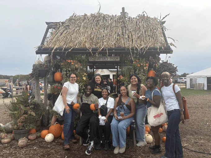 Eight ELPA students sit and stand in front of a festive shack at Schuster's Farm. They are smiling at the camera, with some students holding small pumpkins and some holding fall drinks and treats.
