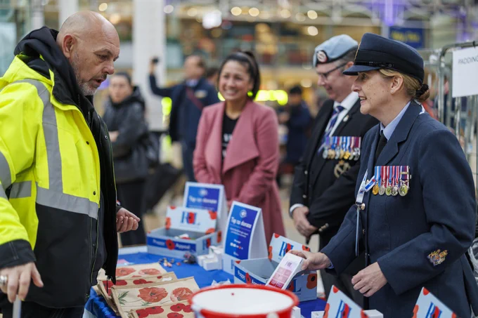 A female RAF personnel is behind a Royal British Legion poppy stand speaking to a member of the public as they buy a poppy for remembrance. Poppies can be seen on the RBL stand.