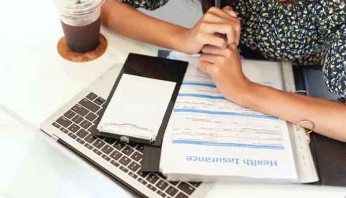 Woman reviewing a Health Insurance Policy document on her laptop, with a notepad and iced coffee nearby — representing financial planning, insurance management, and healthcare coverage decisions.