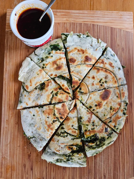 Spring onion pancake served on a wooden chopping board. The Pancake is already cut into eight wedges, and a ramekin of the dipping sauce lays next to it.