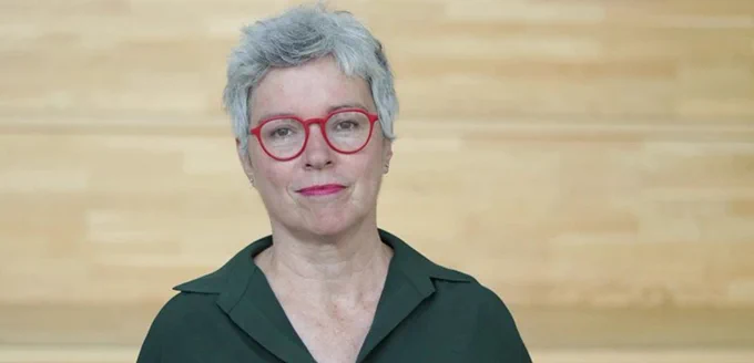 Person with short gray hair and red glasses, wearing a green shirt, standing in front of a wooden background.