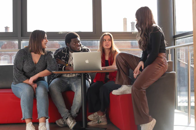 Four individuals engaged in a discussion around a laptop in a well-lit room with large windows.