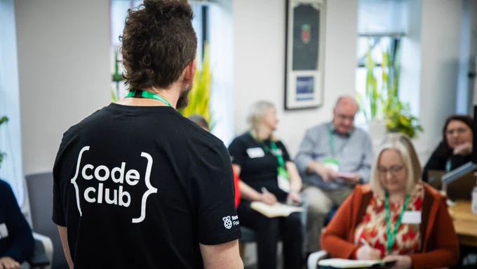 An educator wearing a black Code Club t-shirt leads a group discussion in a bright room with seated participants taking notes.