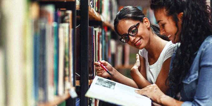 Two women sitting between library shelves, studying a book together with one holding a pencil for note-taking.