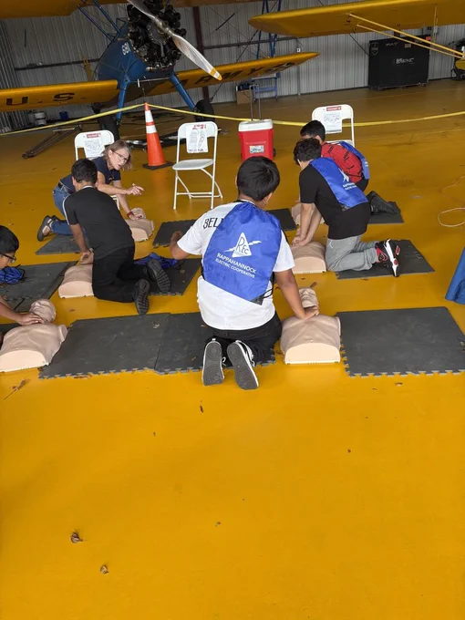 A group of young people, mostly boys, are practicing CPR on mannequins laid out on black mats in what appears to be an airplane hangar with a yellow floor. An adult woman oversees them, and a vintage yellow biplane is visible in the background. Many of the participants are wearing blue vests with "Rappahannock Electric Cooperative" written on them.