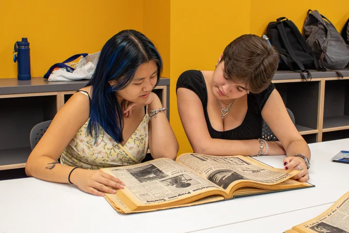 Two people sit at a table and look at a bound copy of a newspaper