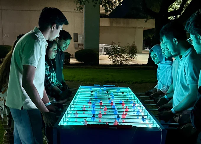 A group of people playing foosball on a brightly illuminated table outdoors at night.