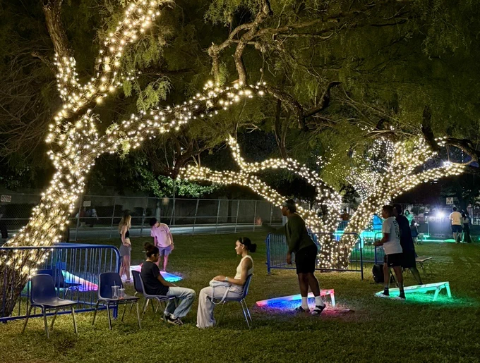 Alt Text: People enjoying a Bestfest, UTSA fall festival, Family zone while playing cornhole under trees beautifully adorned in white string lights.