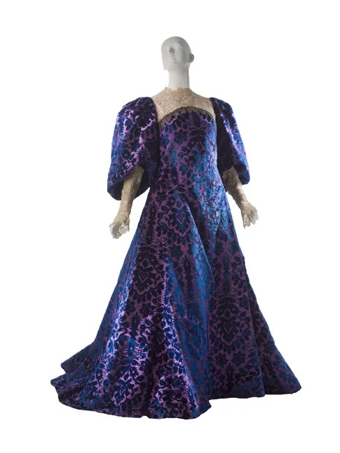 A mannequin wearing a purple voided velvet gown with broad skirt and big sleeves. It has a white lace yoke around the neck