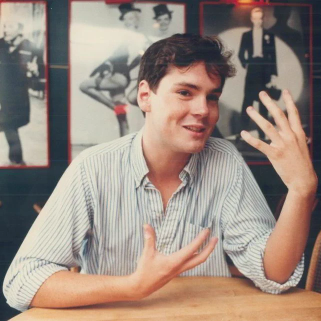 A color photograph of a young man sitting at a wooden table in a café or restaurant, gesturing with both hands. He is wearing a light blue and white striped shirt with rolled-up sleeves. The background features dark walls adorned with framed black-and-white posters of people in various poses, including a dancer and a man in a suit. The setting includes wicker chairs and a warm, dimly lit ambiance.