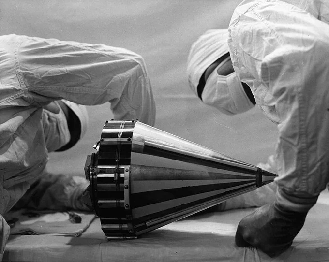 People wearing masks, gloves, and clean room bunny suits while assembling a nose cone containing a scientific instrument.