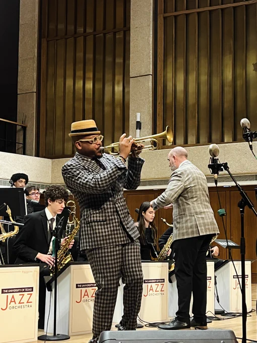 A musician plays a trumpet solo on stage with other band members, including a saxophonist, in front of a sign reading "Jazz Orchestra." The backdrop features large golden panels.