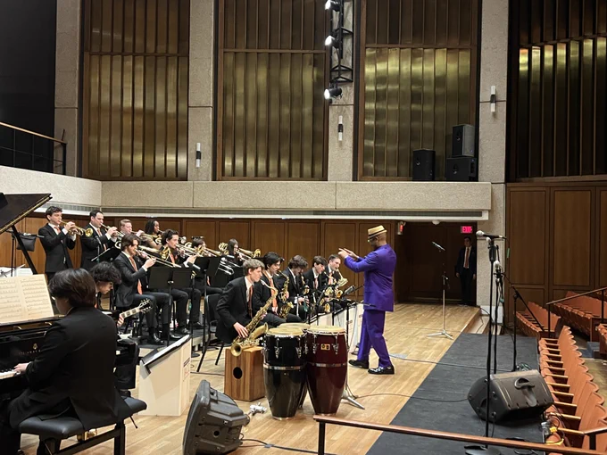 A conductor in a purple suit leads a jazz band performance on stage at a concert hall. The band members are dressed in black and playing various brass instruments and saxophones, with a piano also visible on stage.