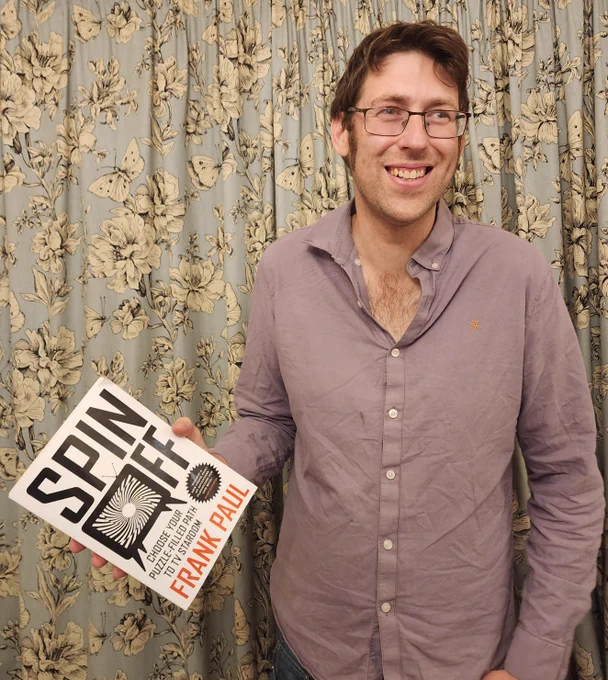 A picture of me smiling and gazing up to my left, wearing a greyish-purple shirt in front of pale blue curtains patterned with cream flowers and butterflies.  I'm holding a copy of my new book, Spin Off, which has a striking cover (the title in thick, angular black letters, with a TV set containing a swirling, hypnotic pattern replacing the "O" of "Off", and my name in red underneath).