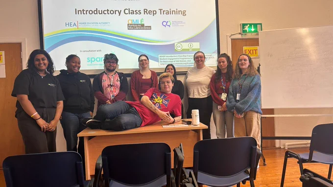 Student trainers Elle and Ebere with class reps in Carlow College after a training session. They are standing in front of a projection screen and smiling.