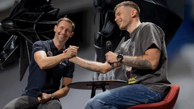 Polish astronaut Sławosz Uznański-Wiśniewski sits on stage with a CD Projekt Red employee. The two smile broadly and lock hands in an embrace. A large screen stands behind them, featuring an image of two medallions from The Witcher floating in outer space aboard the International Space Station.



