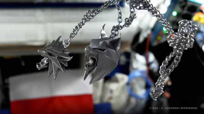 Two medallions from The Witcher, the Wolf and the Lynx, float in outer space aboard the International Space Station. Their chains wrap together in the zero gravity, and in the background is the Polish flag.