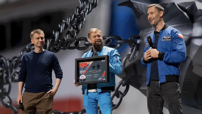 CD Projekt Red Co-CEO Michał Nowakowski and CFO Piotr Nielubowicz stand on stage with Polish astronaut Sławosz Uznański-Wiśniewski, all three of them smiling. Nowakoski and Uznański-Wiśniewski hold microphones while Nielubowicz holds a frame containing the Wolf and Lynx medallions from The Witcher. It’s decorated with images of the two medallions floating aboard the International Space Station and logos and other imagery of the European Space Agency. A large screen featuring an image of the medallions floating in outer space stands behind them.

