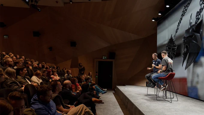 Polish astronaut Sławosz Uznański-Wiśniewski sits on stage with a CD Projekt Red employee, having a conversation as a large audience of CD Projekt Red employees sits listening attentively. A large screen stands behind them, featuring an image of two medallions from The Witcher floating in outer space aboard the International Space Station.

