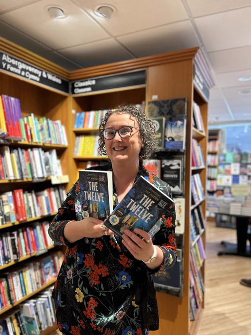 A photo of Liz Hyder, an author, holding up two copies of her book The Twelve and smiling for the camera. She has dark curly hair and is wearing a floral patterned dress.