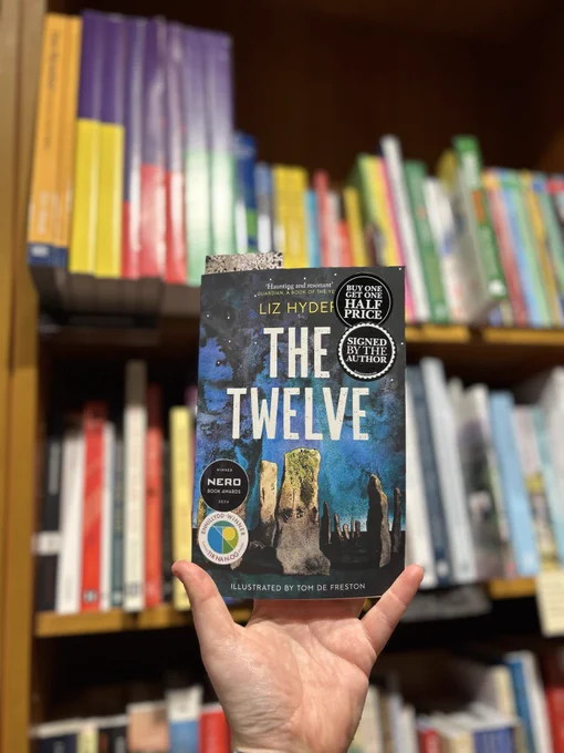 A photo of a white hand holding up a paperback copy of The Twelve by Liz Hyder against a background of bookshelves.