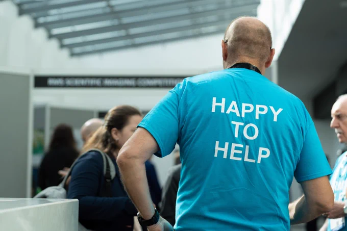 Man wearing a bright blue shirt with 'HAPPY TO HELP' printed on the back at a busy indoor event.
