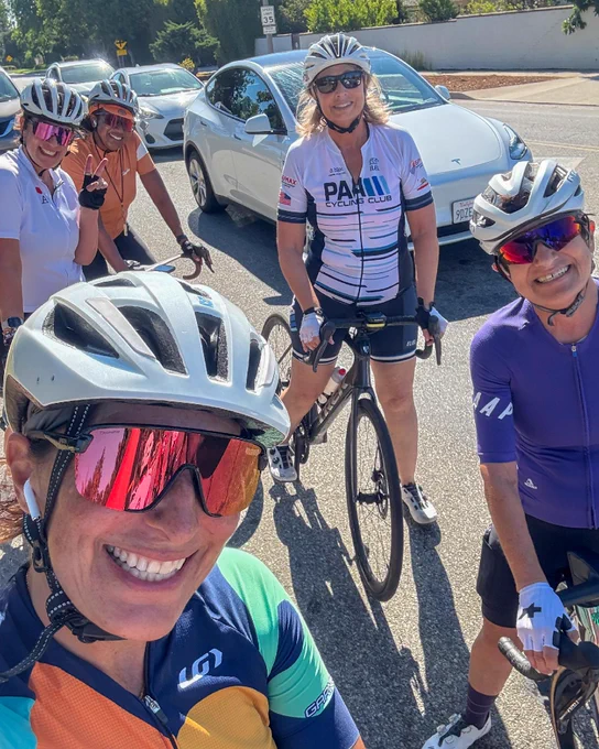Four cyclists in various colorful outfits, including helmets and sunglasses, smiling for a selfie on a sunny day with cars and greenery in the background.