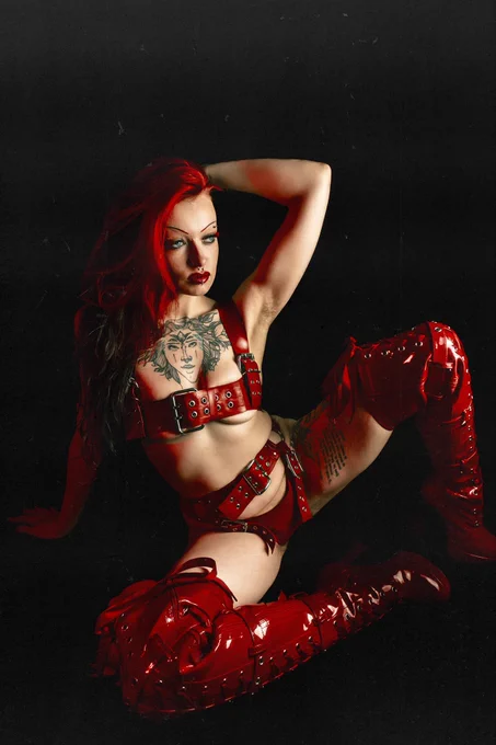 Countess wears shiny red thigh high heeled boots with buckles and laces, and a red latex harness lingerie set. Sitting on the floor leaning back on one hand, with one knee raised and their hand in their red and black hair which is swept to one side, with her blue eyes looking off camera.